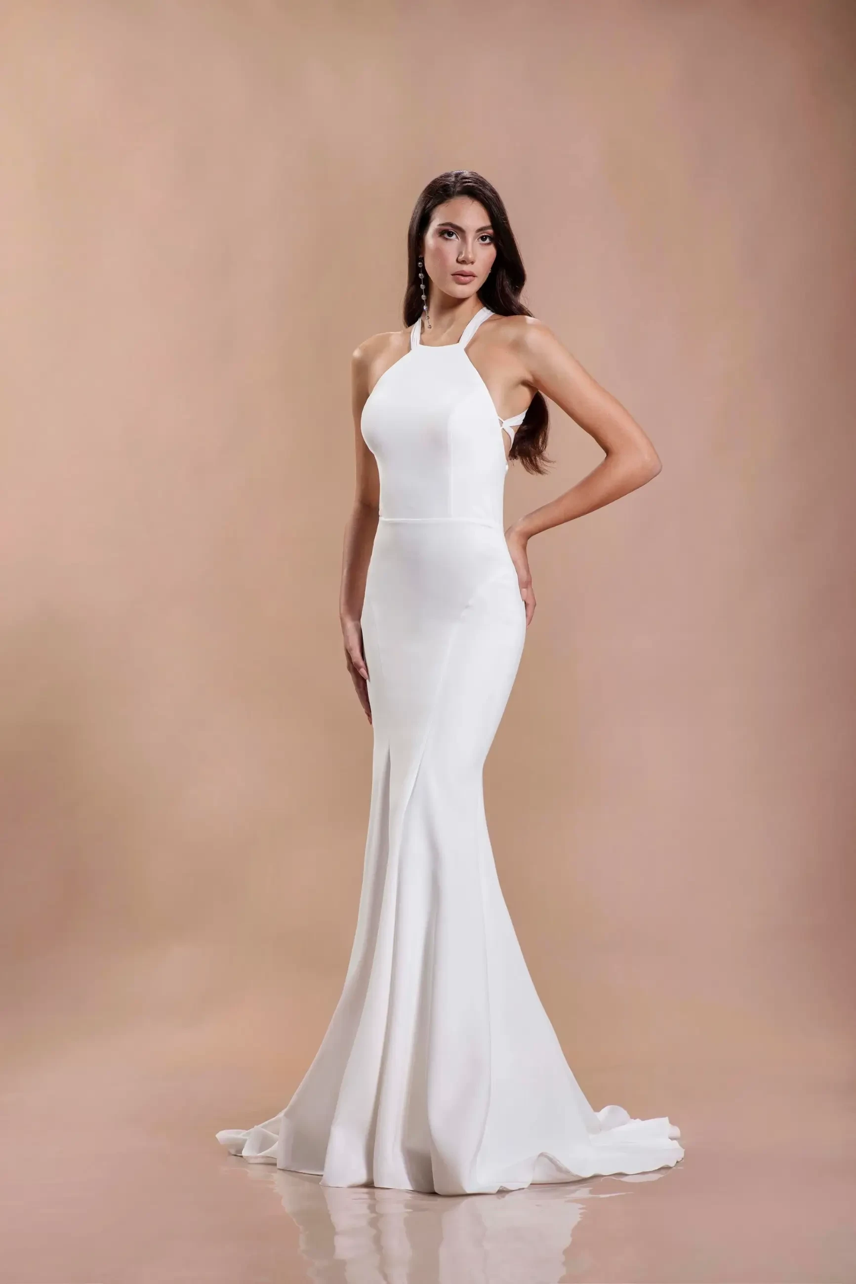 A woman in a sleek, floor-length white gown poses against a soft brown backdrop. The dress features a halter neckline and a mermaid silhouette, conveying elegance.