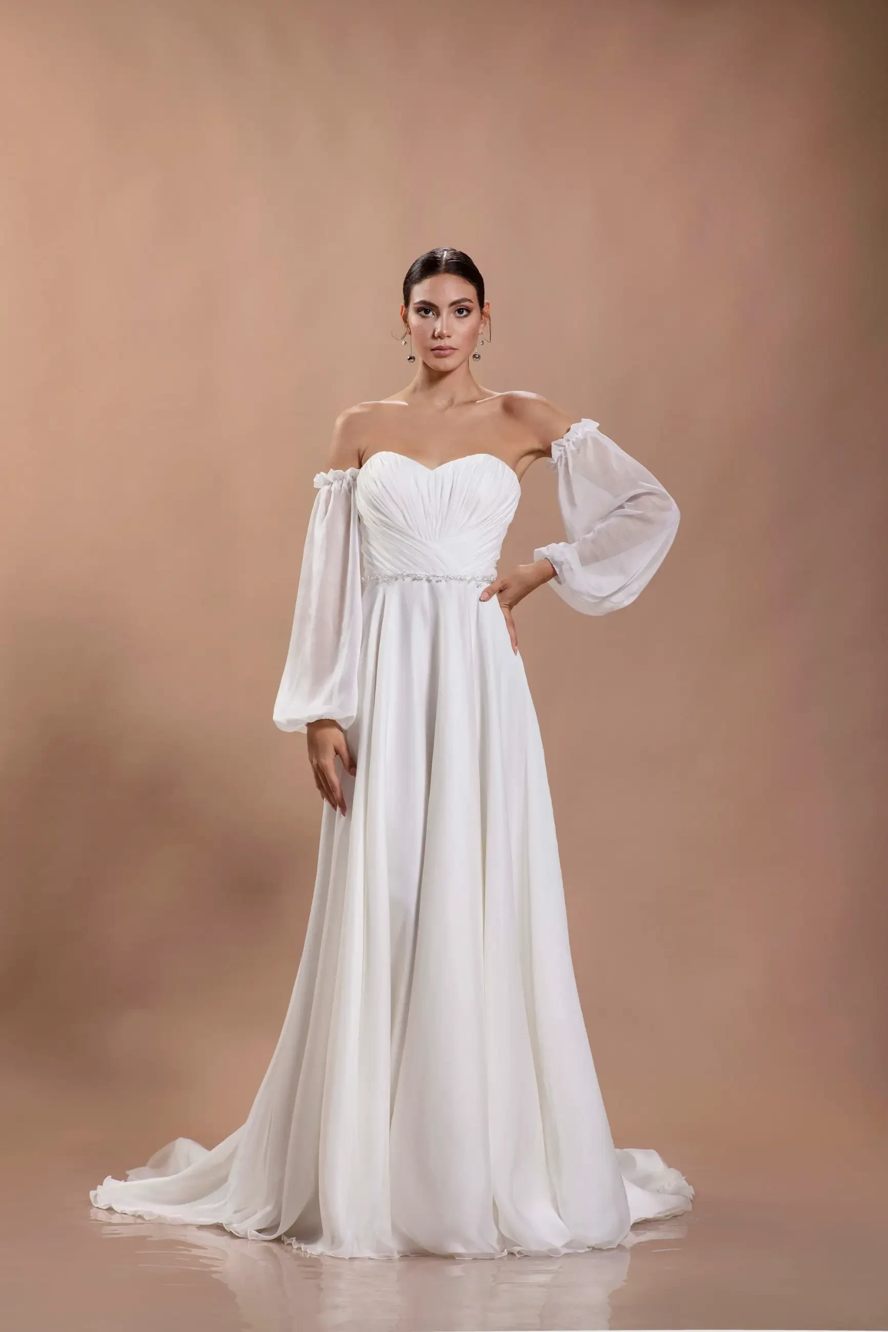 A woman stands confidently in an off-the-shoulder, long-sleeved white wedding gown against a warm, neutral background, exuding elegance and grace.
