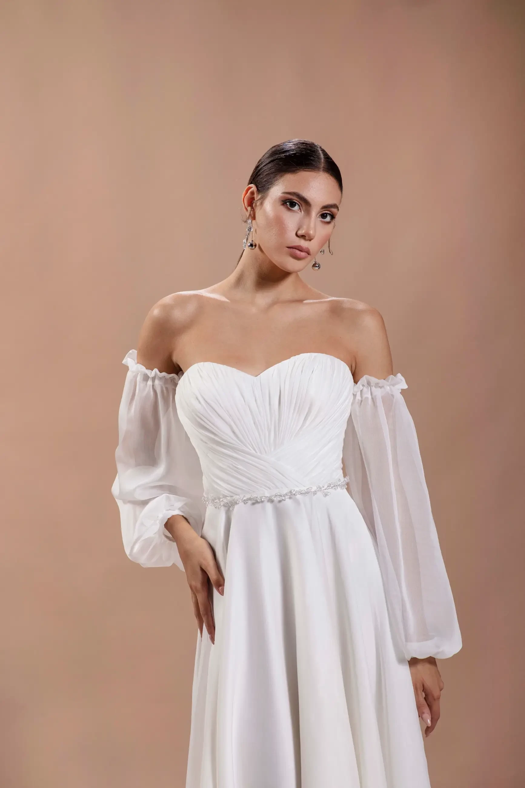 Woman in an elegant off-shoulder white gown with sheer sleeves against a neutral background, exuding a sophisticated and serene expression.