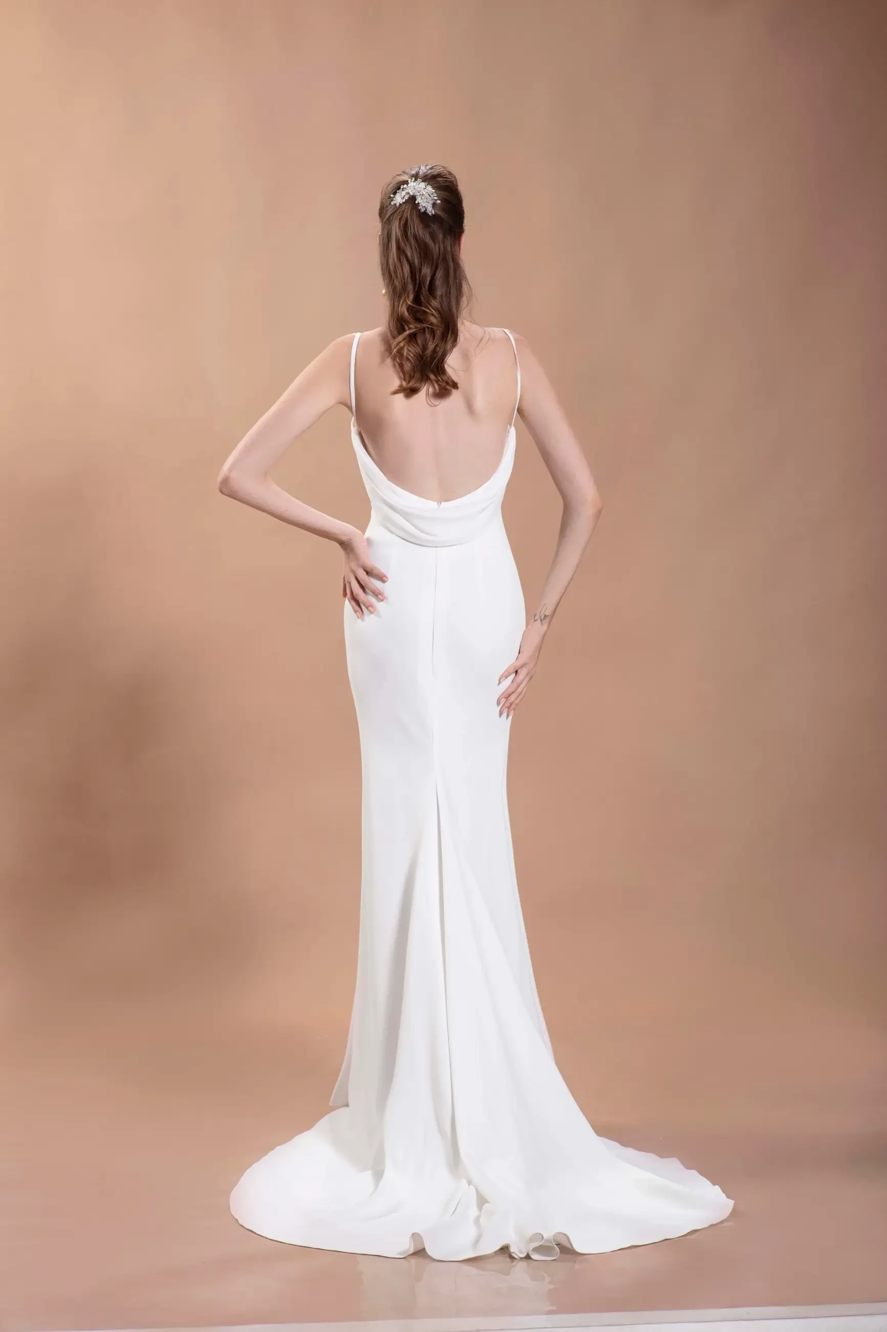 A woman in a white, backless gown with thin straps stands against a soft beige background. Her hair is styled with an elegant accessory.