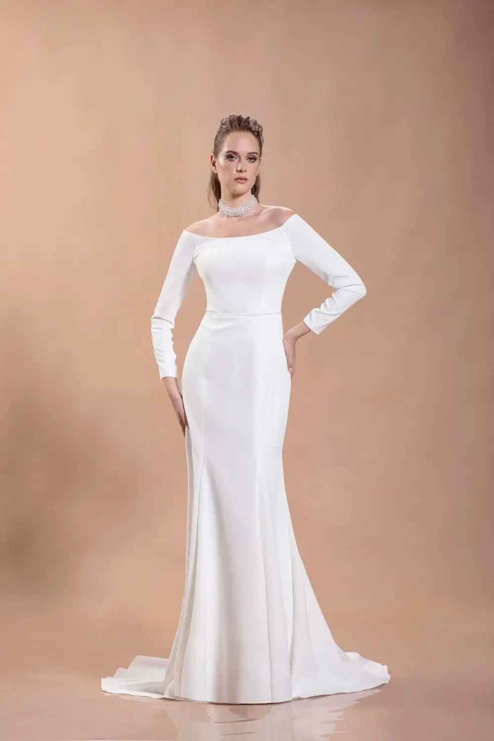 A model wearing a long-sleeved, off-the-shoulder white wedding dress, posing on a beige background.