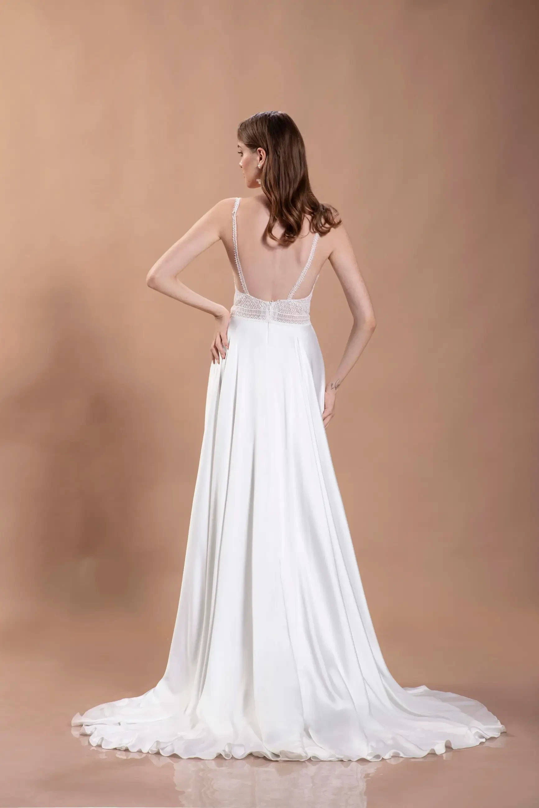 A woman in a flowing white wedding gown with a deep V-back and beaded straps stands against a soft brown background, exuding elegance and grace.