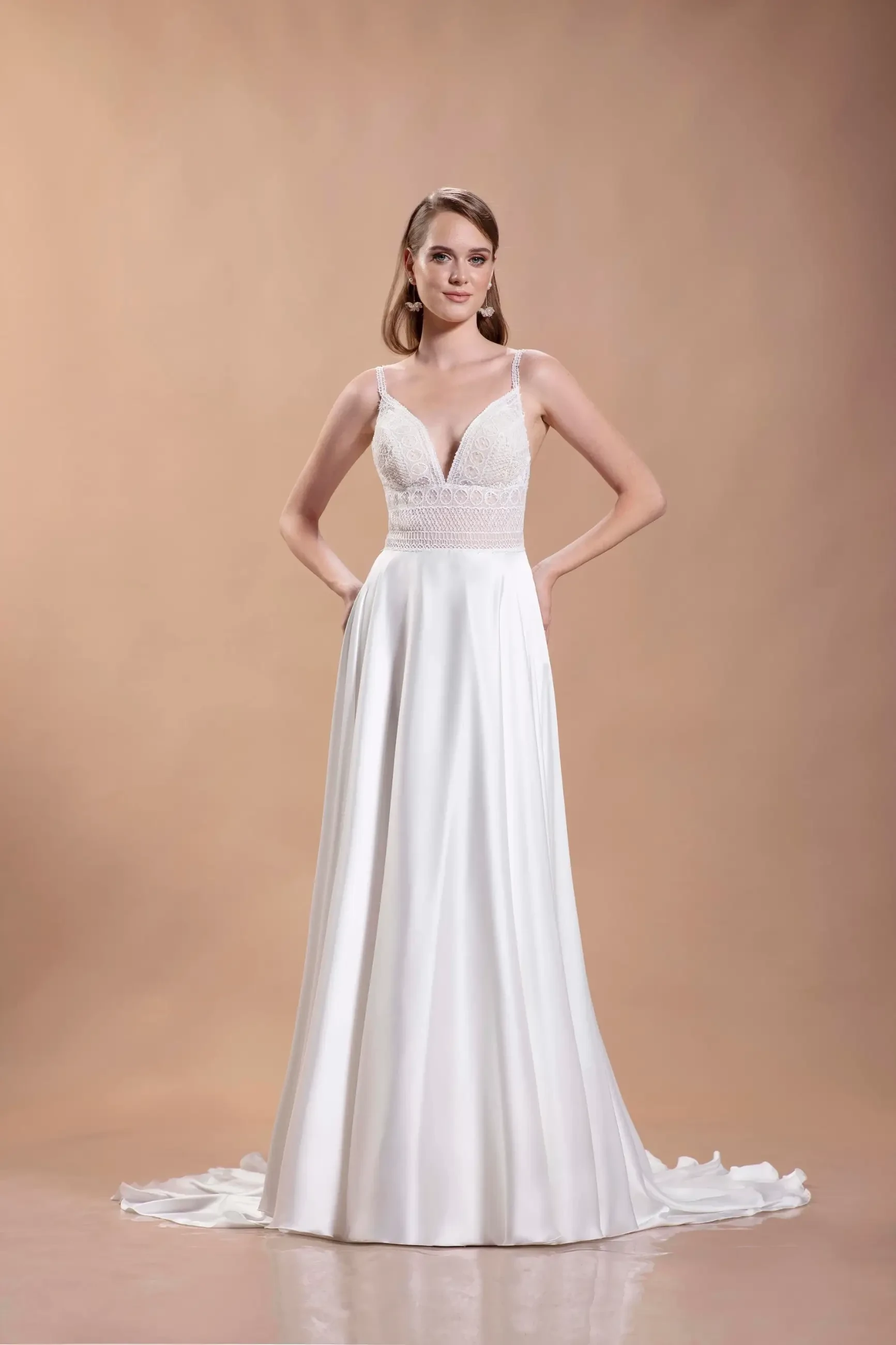 A woman in a sleek, elegant white wedding dress with a lace bodice and flowing skirt, standing confidently against a soft beige background.