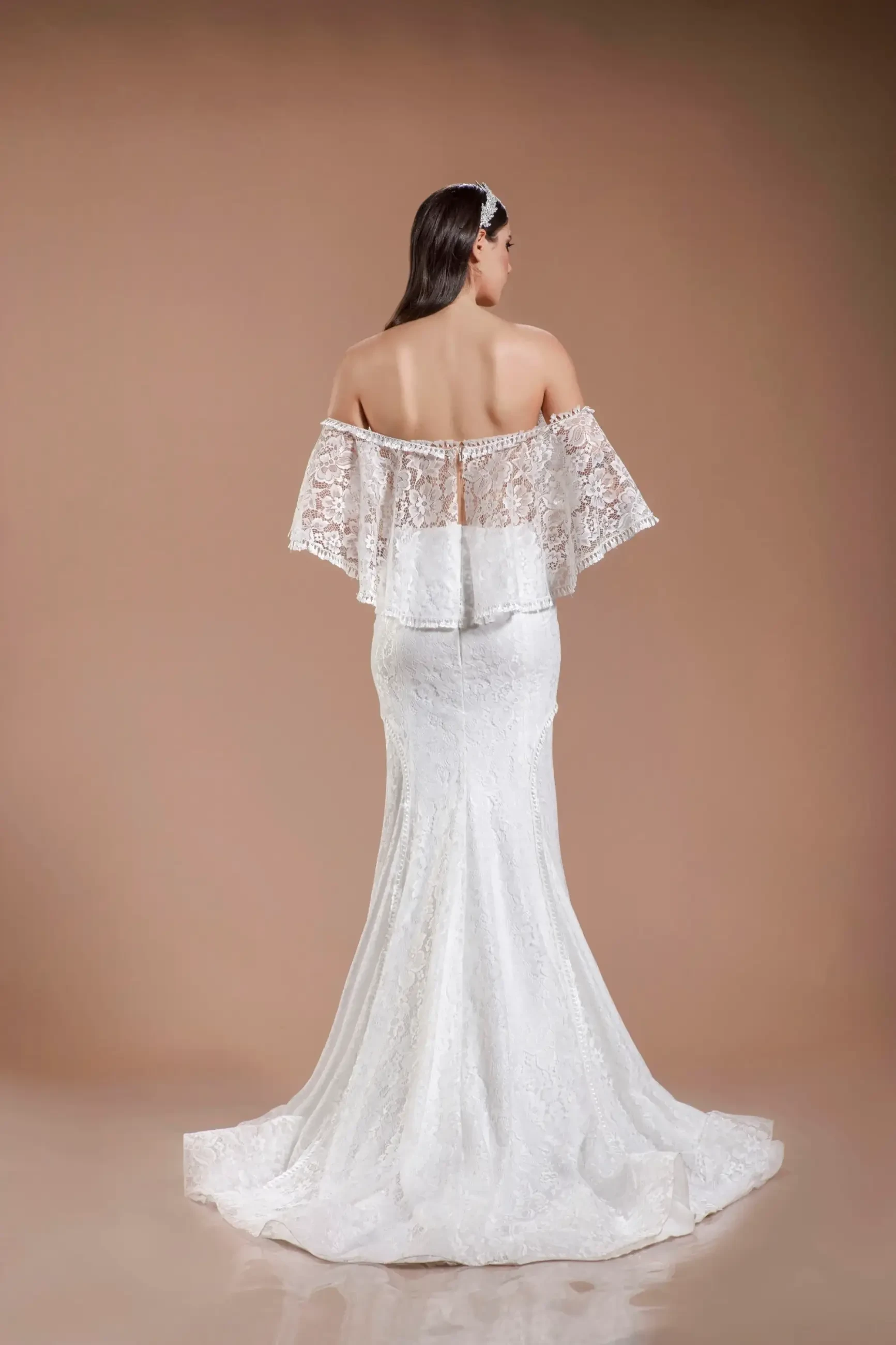 Model is wearing an off-the-shoulder fit-and-flare gown with a flowing lace cape that drapes elegantly behind her.