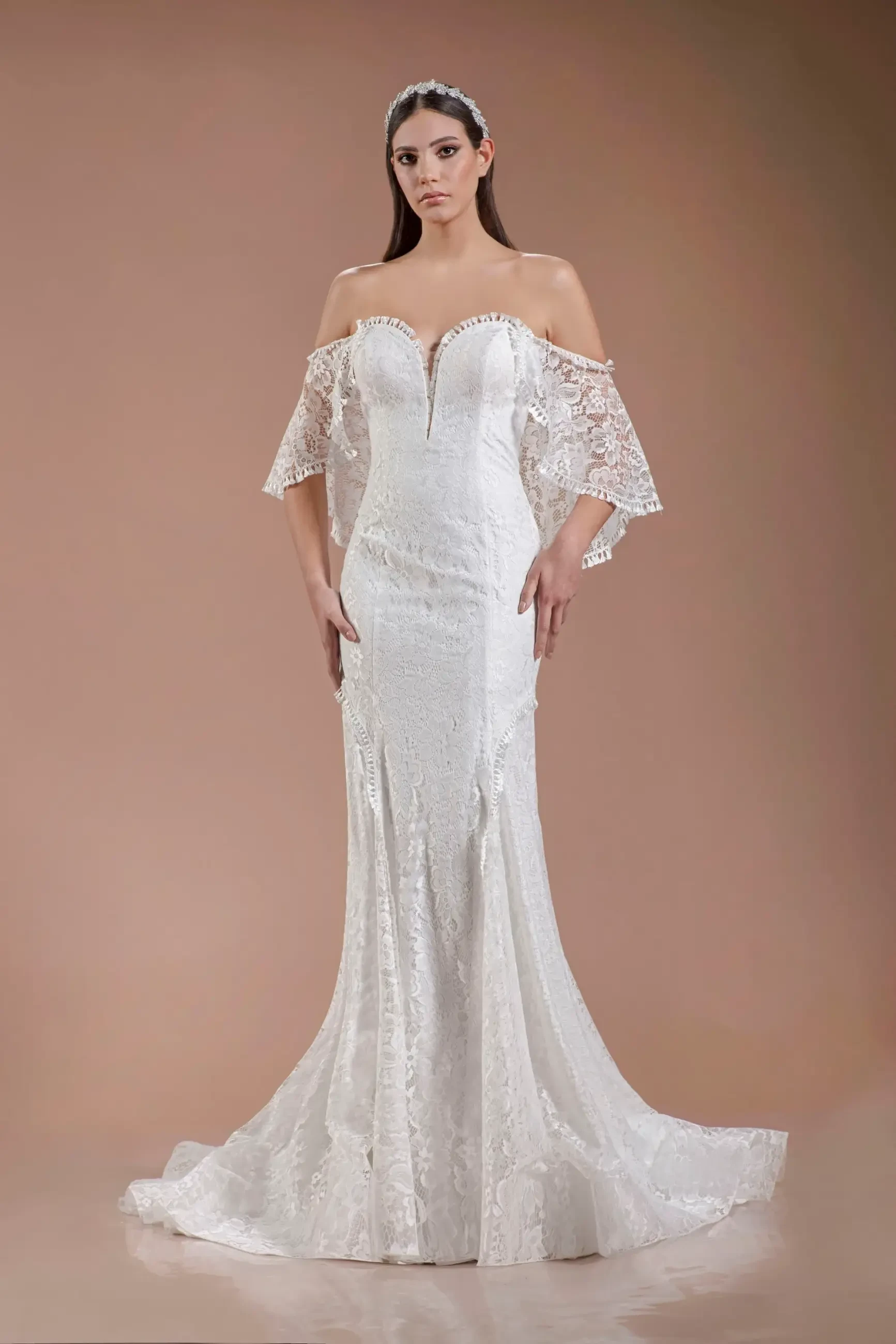 Model is wearing an off-the-shoulder fit-and-flare gown with a flowing lace cape that drapes elegantly behind her.
