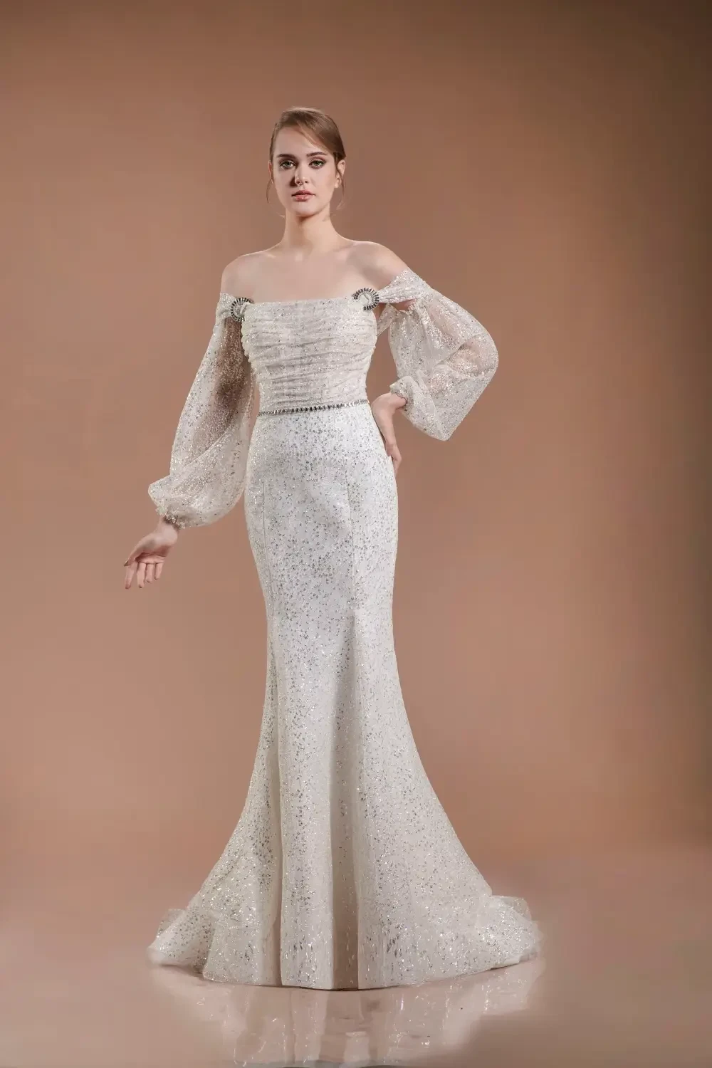 A model wearing an elegant, sparkling white gown with off-the-shoulder sleeves and a fitted silhouette, standing against a neutral background.