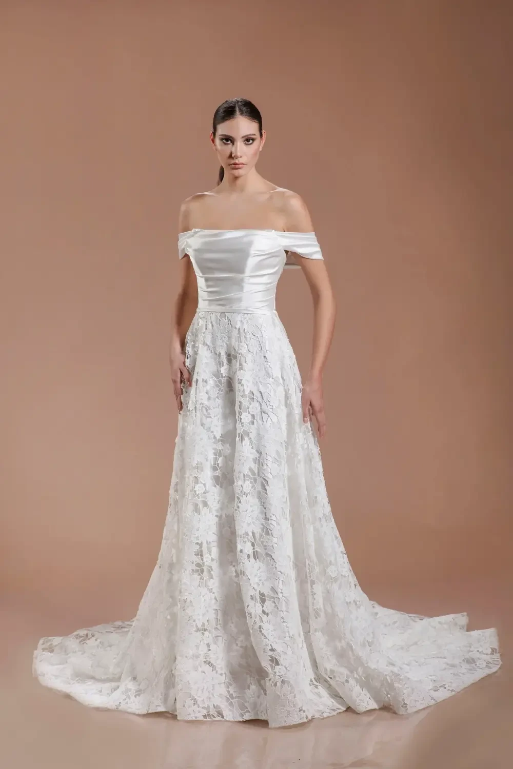 A model in a white off-the-shoulder wedding dress with a lace overlay, standing against a neutral background.