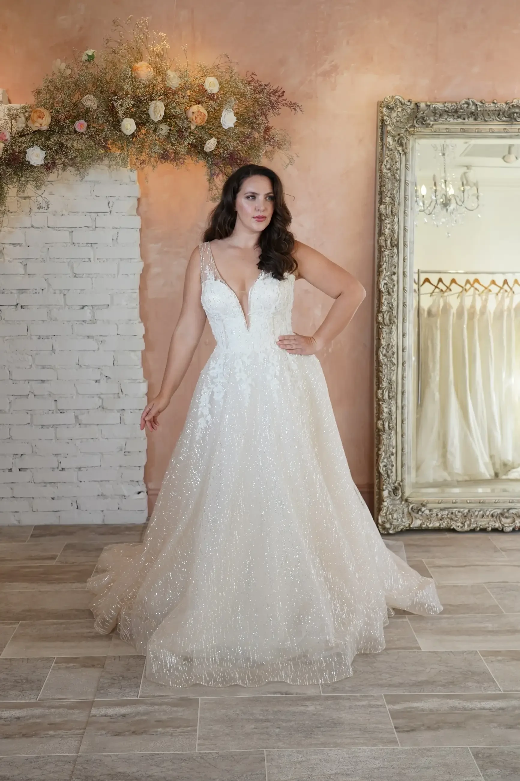 A woman poses in an elegant, shimmering white wedding dress with a deep V-neckline and a full skirt, standing in a beautifully decorated room with floral arrangements and an ornate mirror.