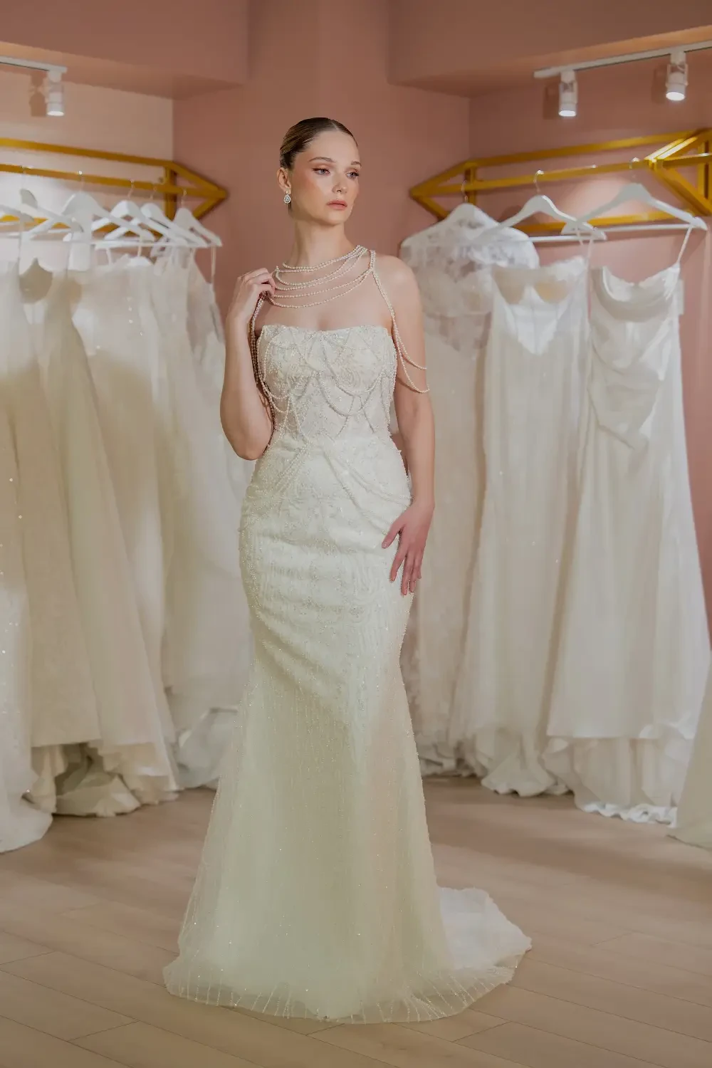 A model in a bridal gown stands elegantly in front of a rack of wedding dresses, showcasing a sophisticated and intricate design.