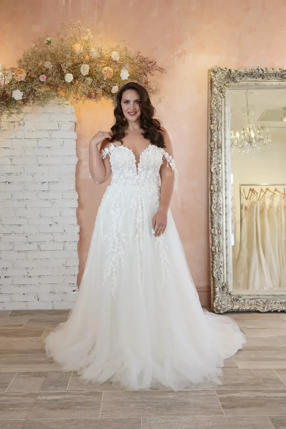 A woman in an elegant, white wedding gown with floral details, standing in a bright room with a brick wall and a large mirror.
