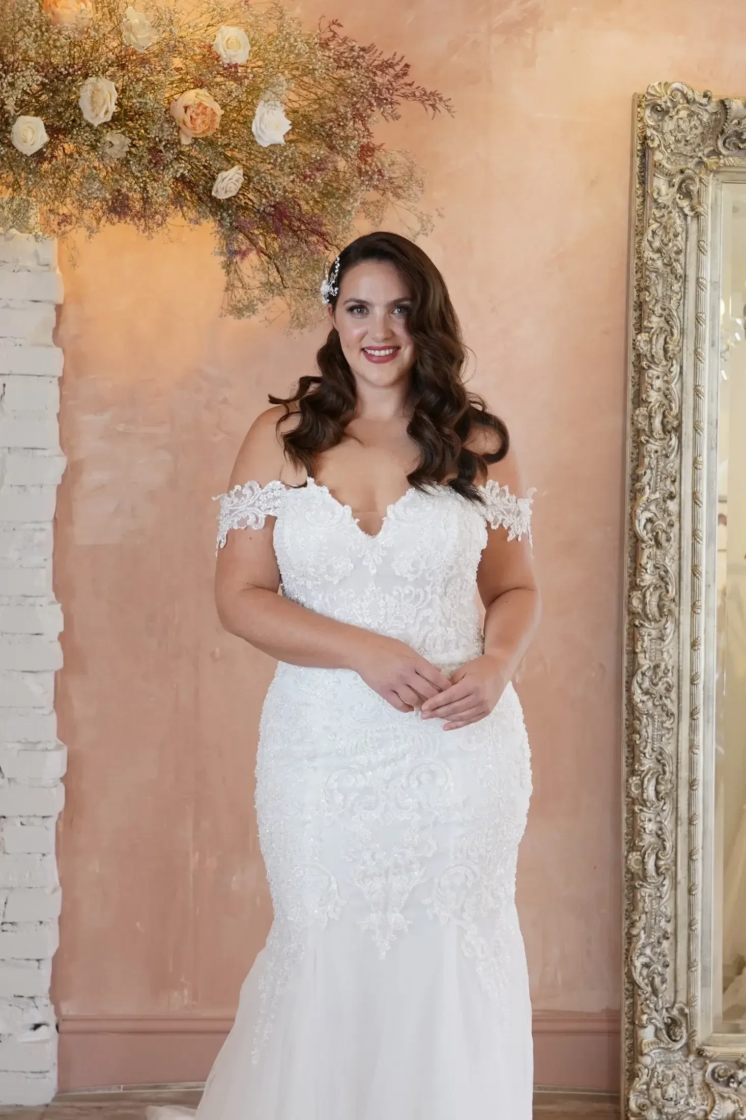 A woman in a wedding dress stands smiling, with wavy hair and a flower accessory, against a softly lit background featuring floral decor and a large mirror.
