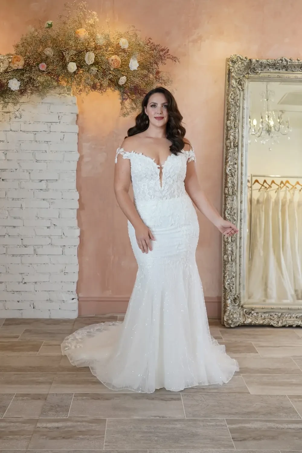 A woman in a white wedding dress stands in a boutique with a floral arrangement and a mirror in the background.