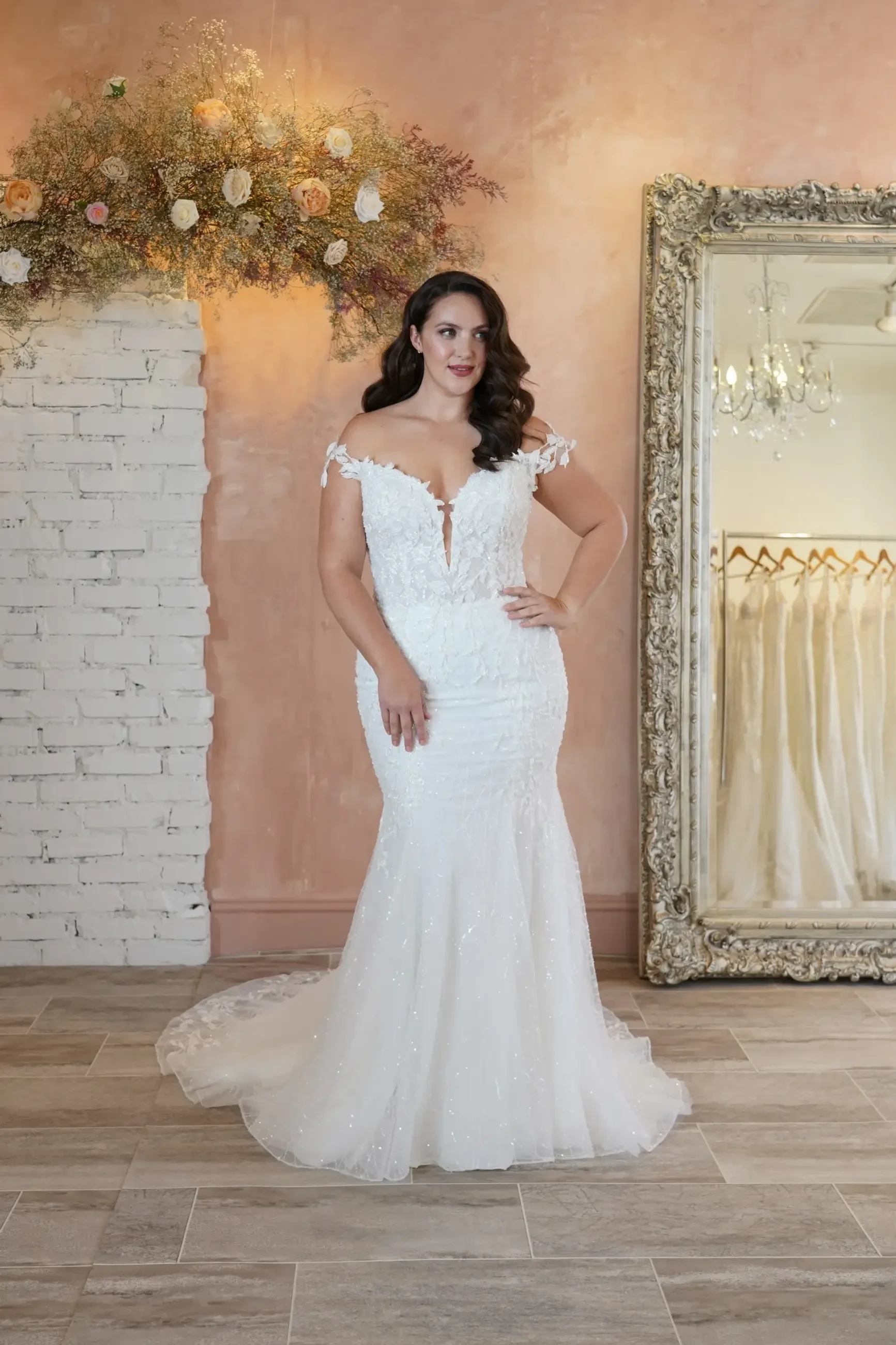 Model is wearing a fit and flare off-the-shoulder wedding gown with a plunging neckline and floral vine lace detailing.