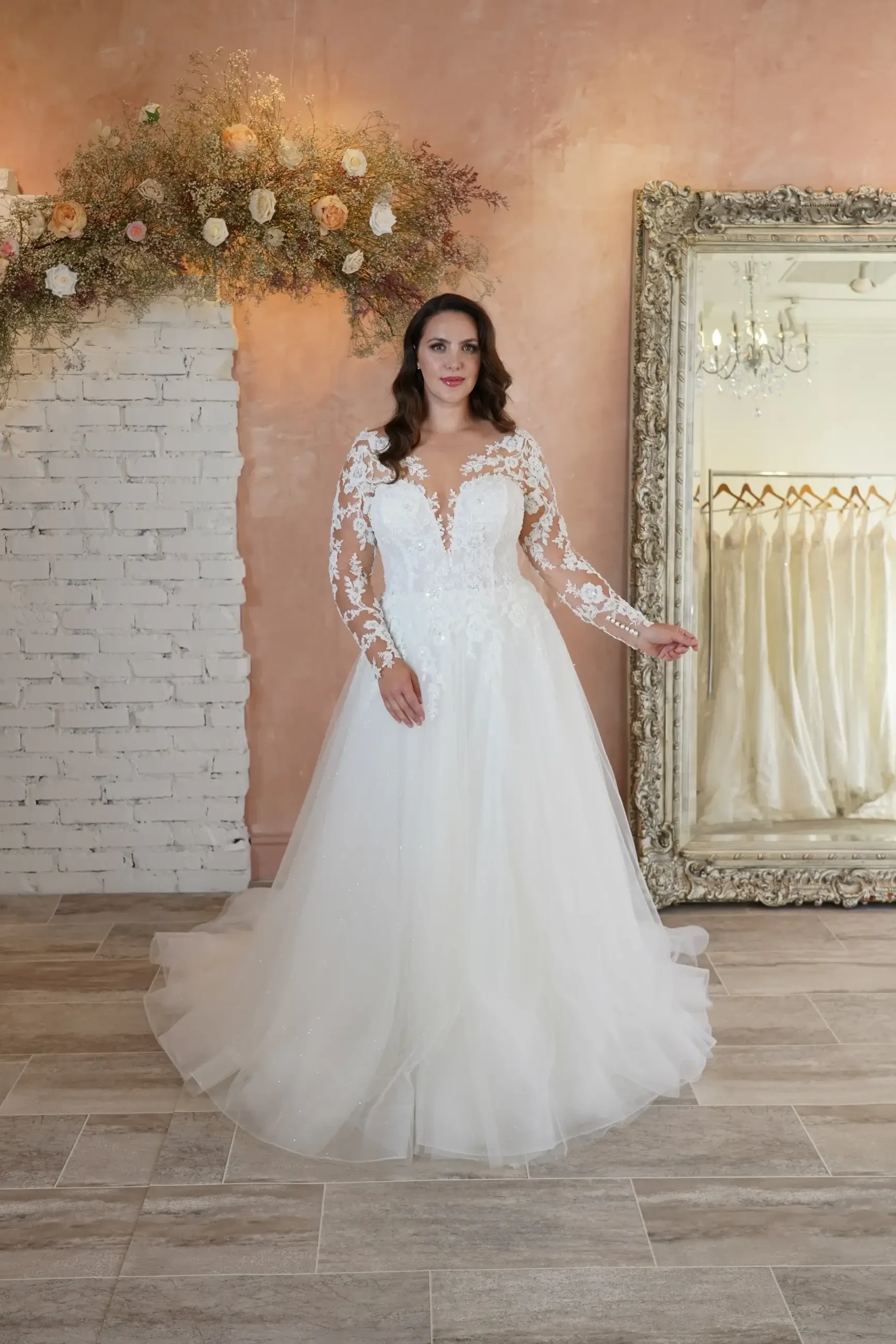 A woman in a white wedding dress with long lace sleeves standing in a bright room with a floral arrangement above her and an ornate mirror.