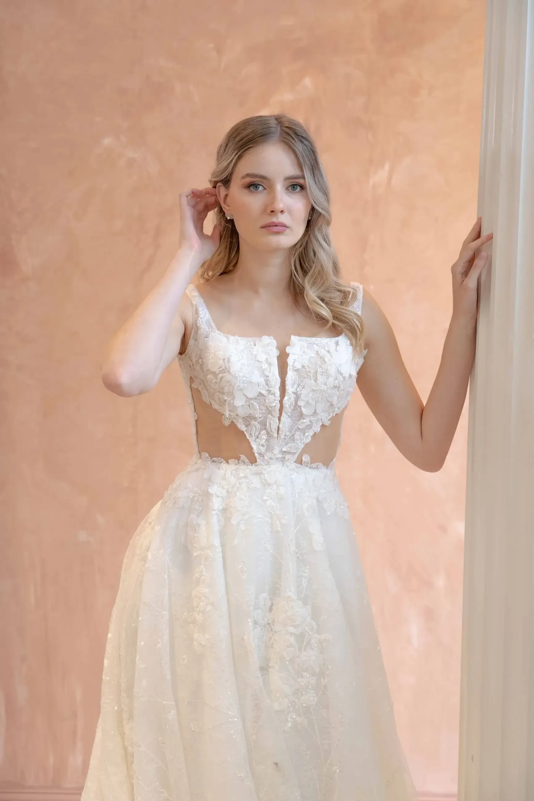 A woman in a detailed lace wedding dress leans against a peach wall, looking serene. Her long hair is styled in loose waves, adding to the elegant tone.