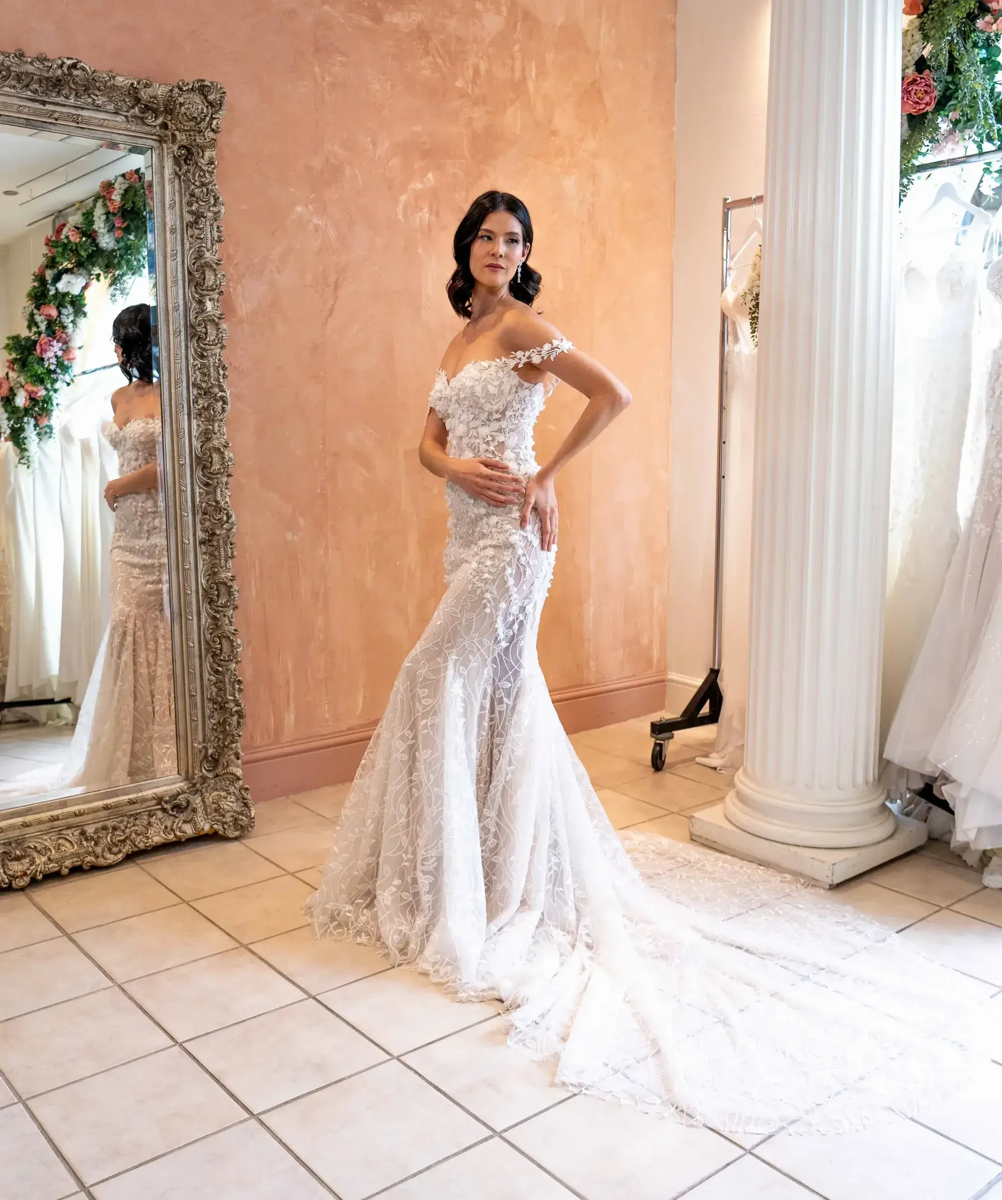 A bride stands elegantly in a fitted lace wedding gown with a long train in a bridal shop. She is near a large ornate mirror, evoking a serene and joyful ambiance.