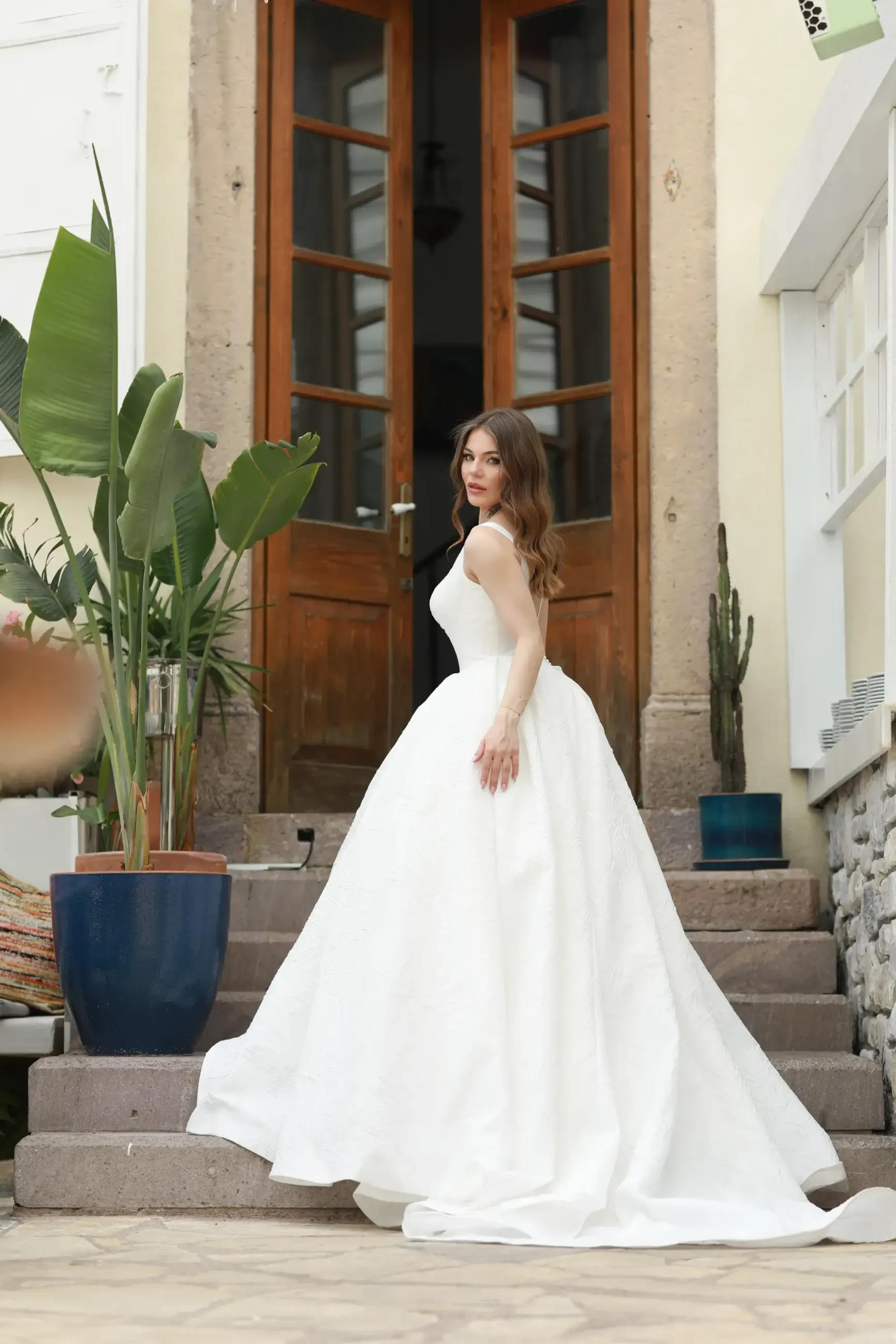 A woman wearing a white wedding dress stands on stone steps in front of a wooden door, surrounded by potted plants.