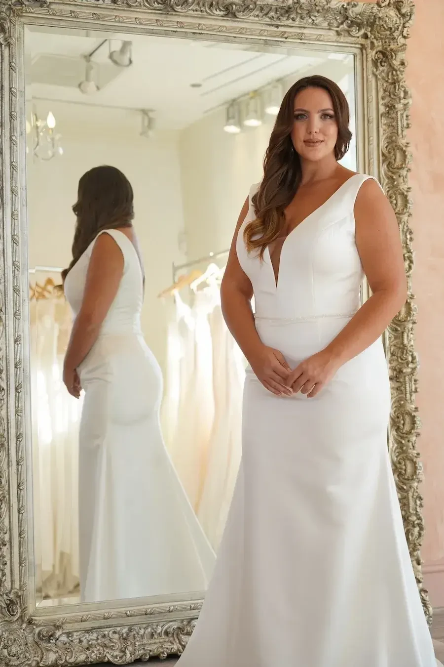 A bride in a white wedding dress stands in front of a large ornate mirror, showcasing her reflection.