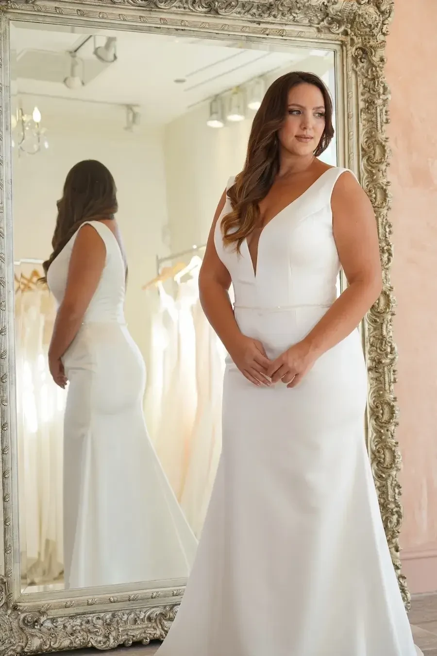 A woman in a white wedding dress stands in front of an ornate mirror, displaying the back of her gown.