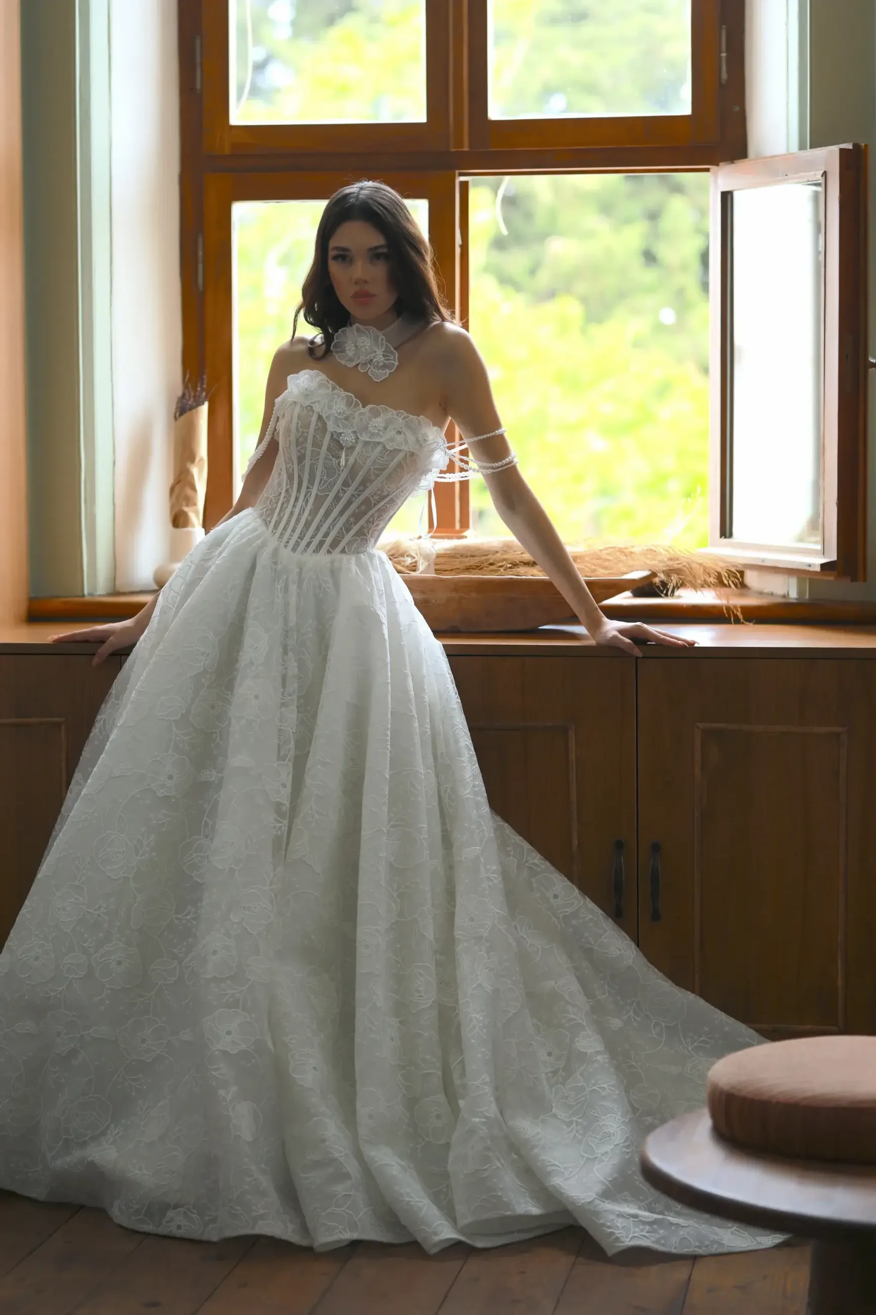 Model is wearing a ballgown with a sculpted sheer bodice, exposed boning, embroidered layers, double straps, and a detachable flower detail at the neckline.