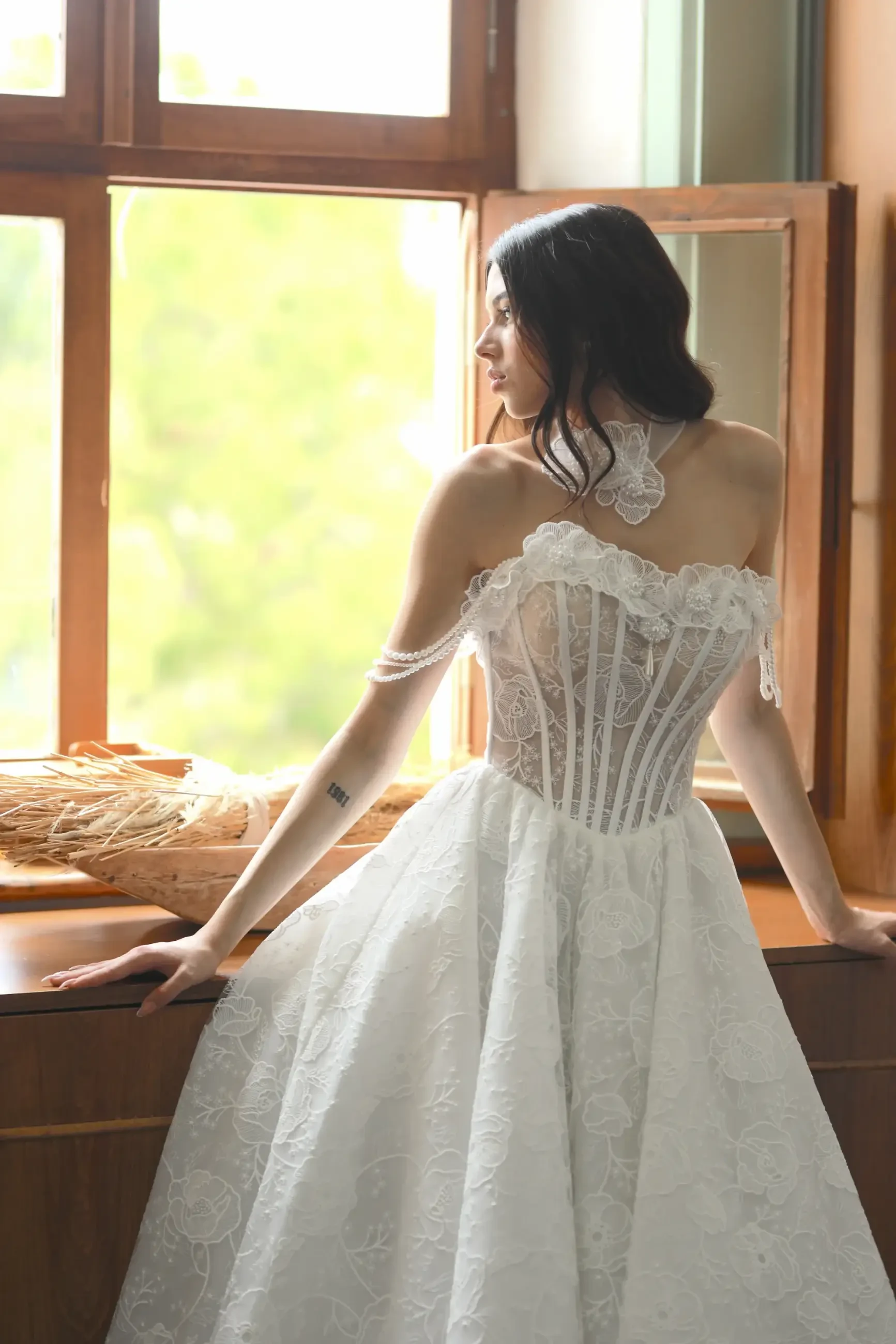 Model is wearing a ballgown with a sculpted sheer bodice, exposed boning, embroidered layers, double straps, and a detachable flower detail at the neckline.