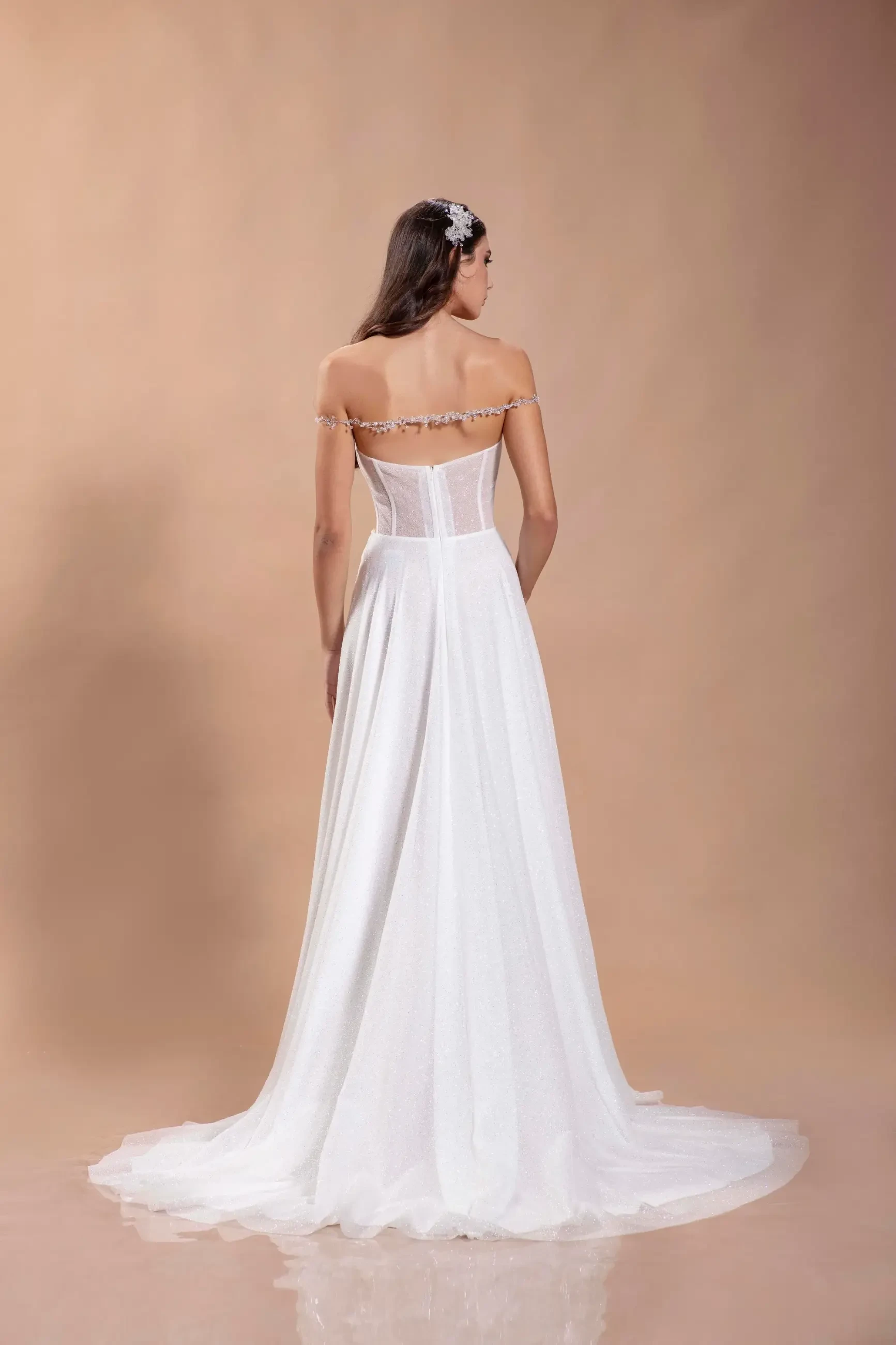 Back view of a woman in an elegant off-the-shoulder white gown with a flowing train. Her hair is styled with a floral accessory. Soft, neutral background.