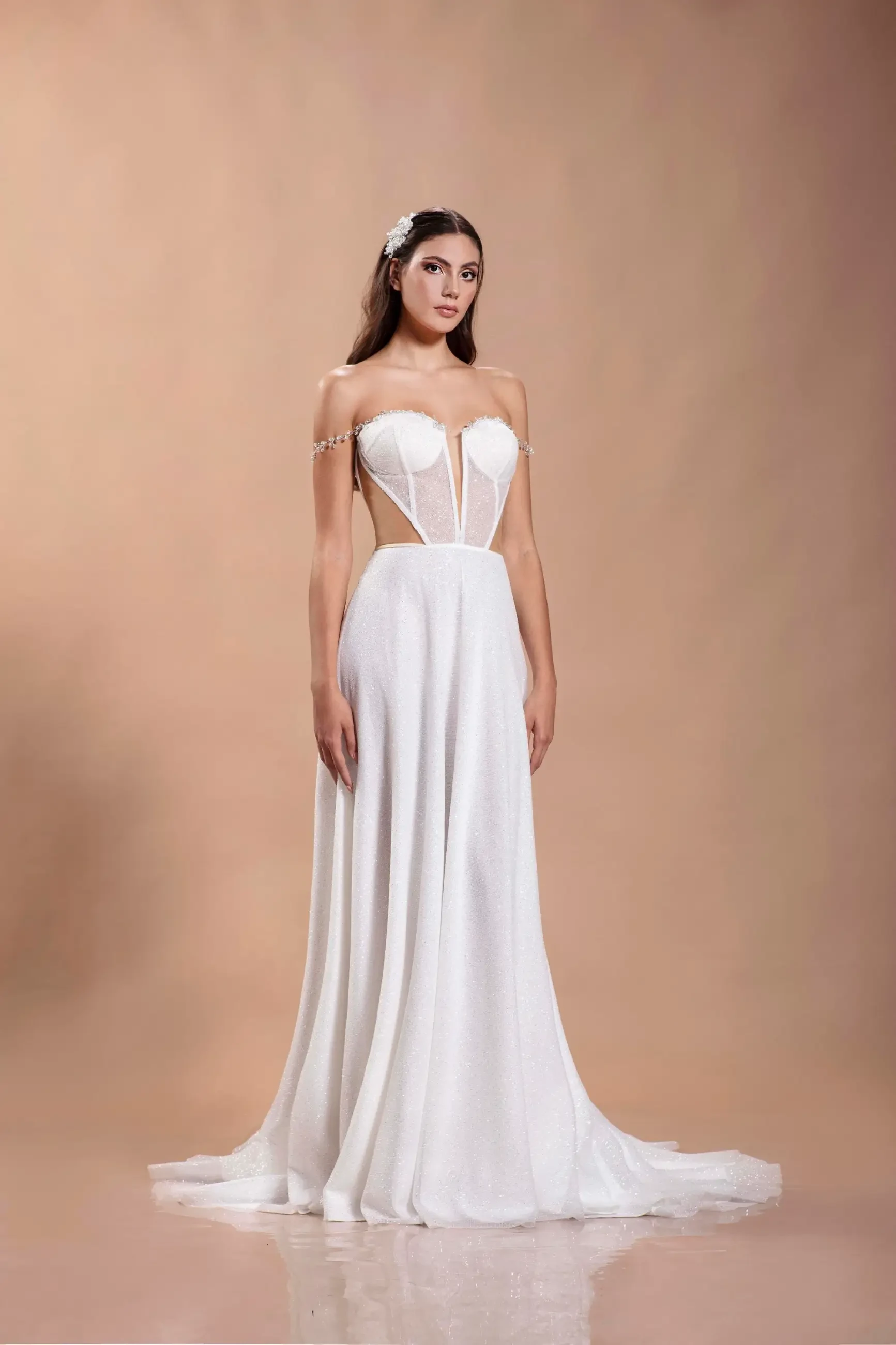 A woman stands gracefully in a strapless white gown with a flowing skirt and sheer cutouts. Her expression is serene, and the background is a soft beige.