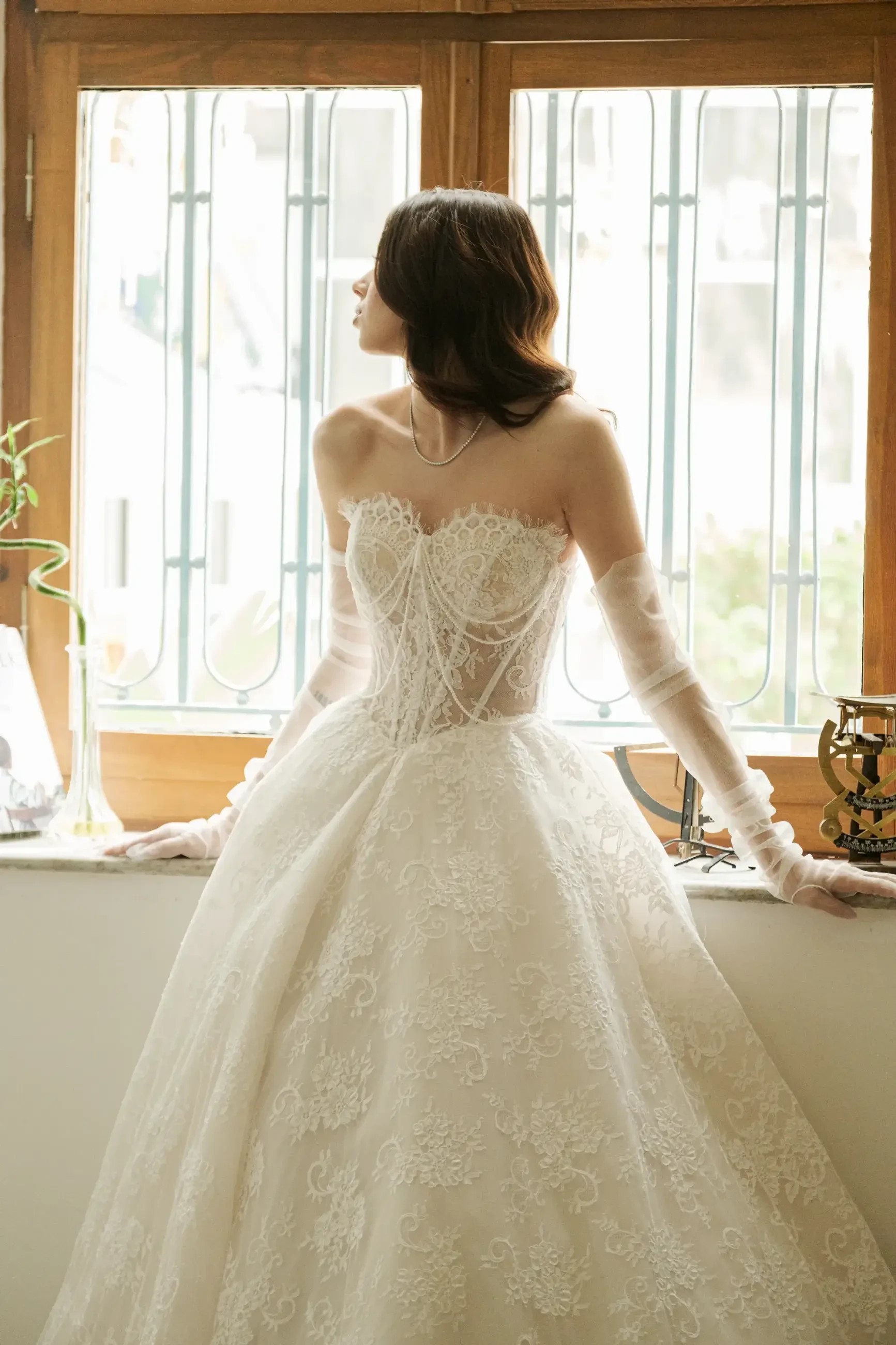 Model is wearing a structured corset bodice gown with lace appliqué and pearl-threaded details, a voluminous tulle skirt, and sheer ruched gloves.