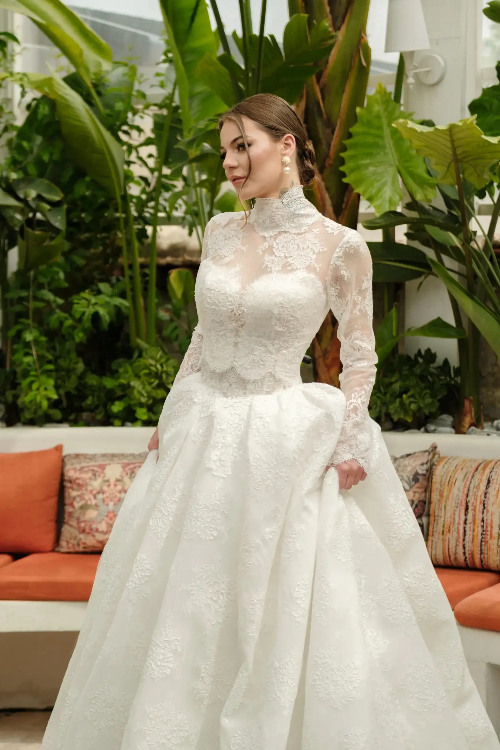 A woman in a long-sleeve, lace wedding dress stands elegantly among tropical plants and a cozy seating area.